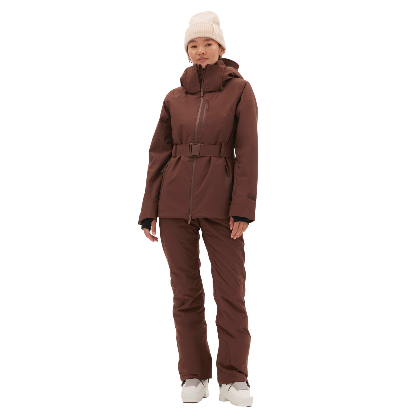 A person stands against a white background, wearing the performance-driven brown Aston Jacket by Halfdays, along with winter pants and a beanie. The look is completed with a belted ski jacket, ready for the mountain's call.