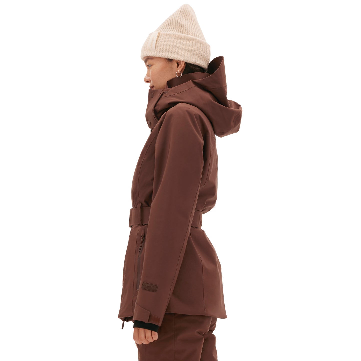 Wearing a belted Aston Jacket by Halfdays with a hood and a white beanie, a person stands in profile against a plain background, prepared for their mountain adventure.