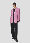 Dressed in Seven Gauge's Double Breast Blazer Tropical Wool in a pink hue, paired with a sleek black shirt and wide-leg pants, as well as matching shoes, a person exudes confidence against a plain background.