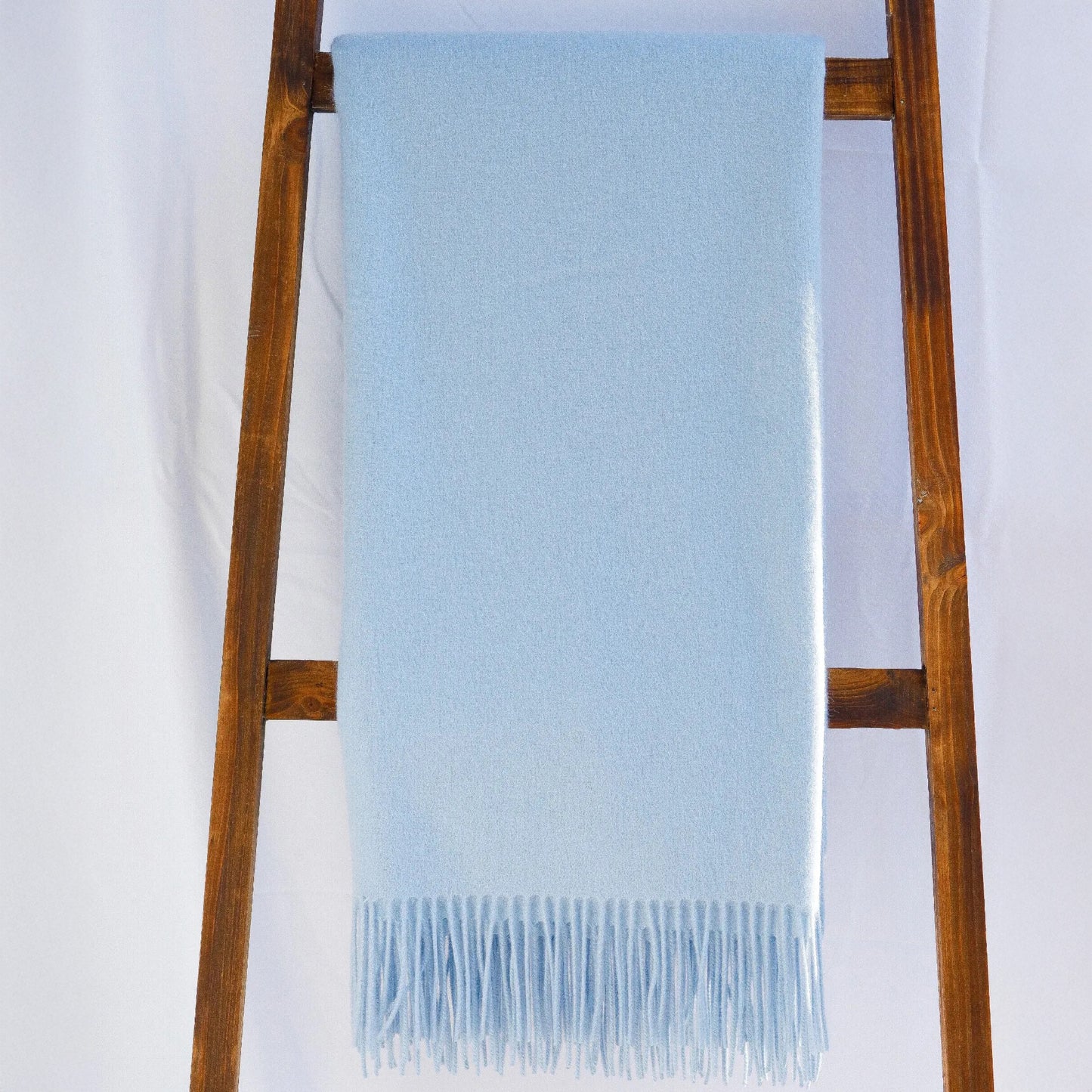 A light blue fringed blanket, The Classic Throw by Alashan, crafted from soft Australian merino wool, draped over a wooden ladder.