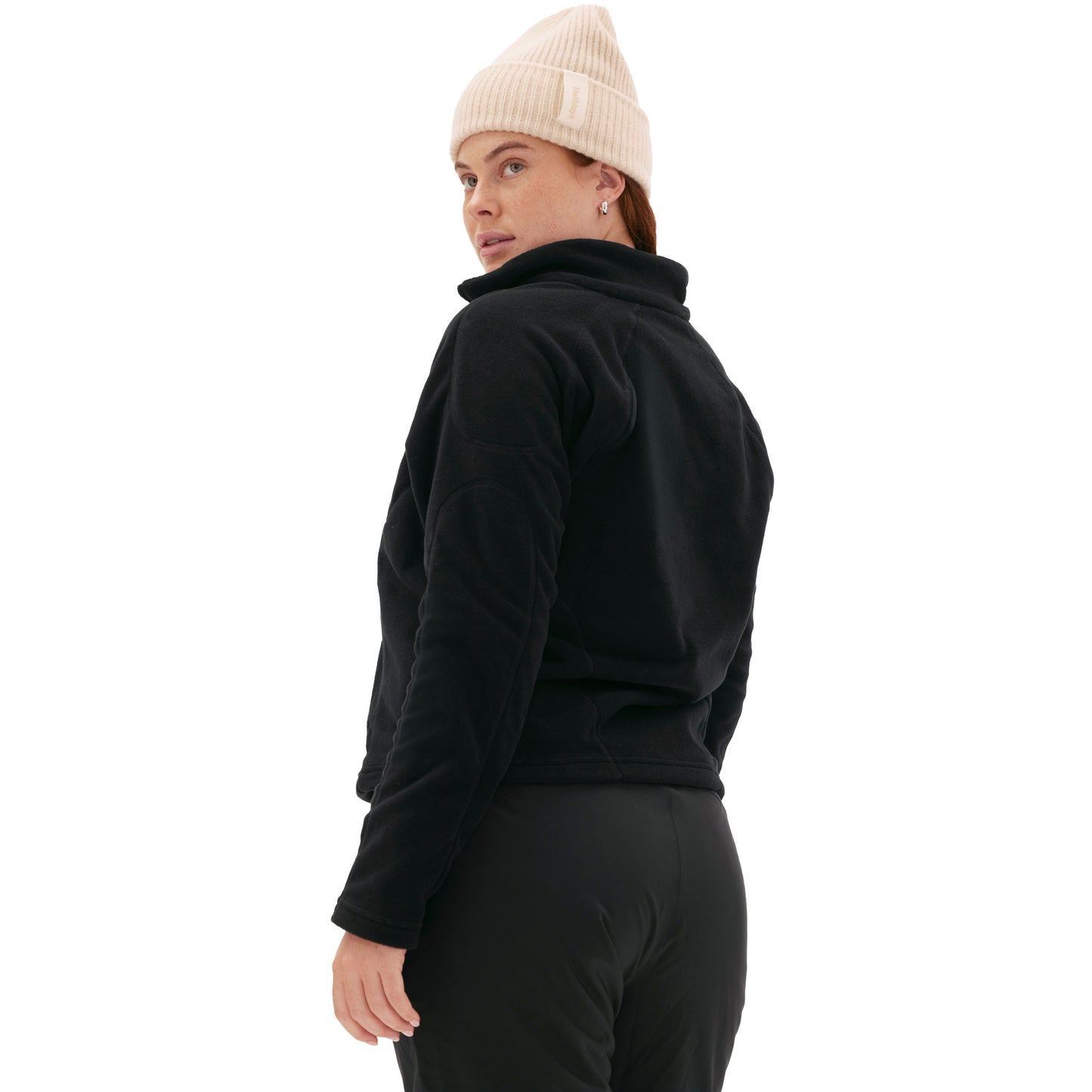 A person wearing the Bonnie Fleece Top by Halfdays and a beige beanie, evoking a high-alpine classic look, gazes back over their shoulder against a white background.