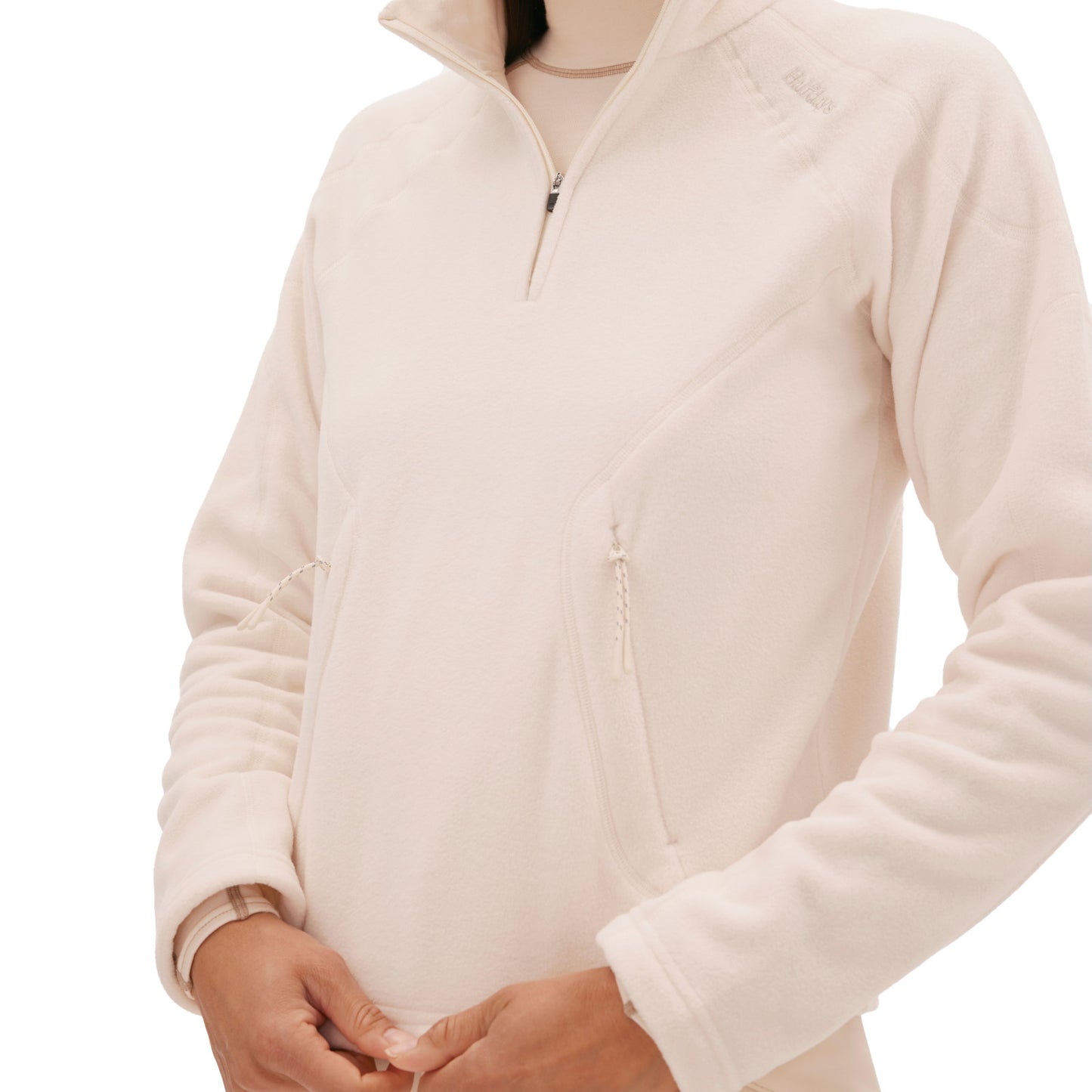 A person is wearing a light beige Bonnie Fleece Top by Halfdays, featuring a quarter-zip Polartec design, with their hands near the front against a white background. Ideal as a performance mid layer, this high-alpine classic combines style and functionality seamlessly.