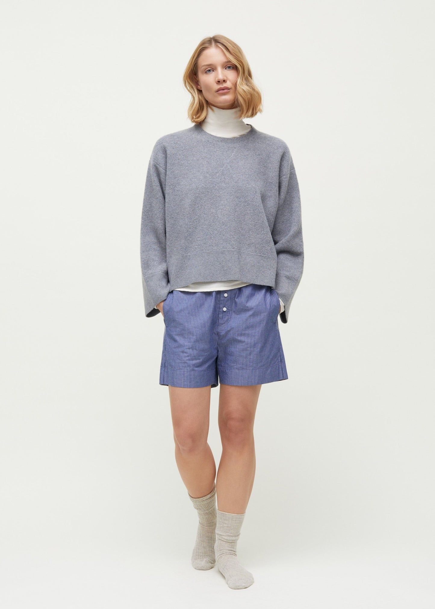 A person stands against a plain backdrop wearing a gray sweater made from 100% Organic Chetna Cotton over a white turtleneck, Aiayu's Letto Casual Shorts in blue stripes, and gray socks. They have shoulder-length blond hair.