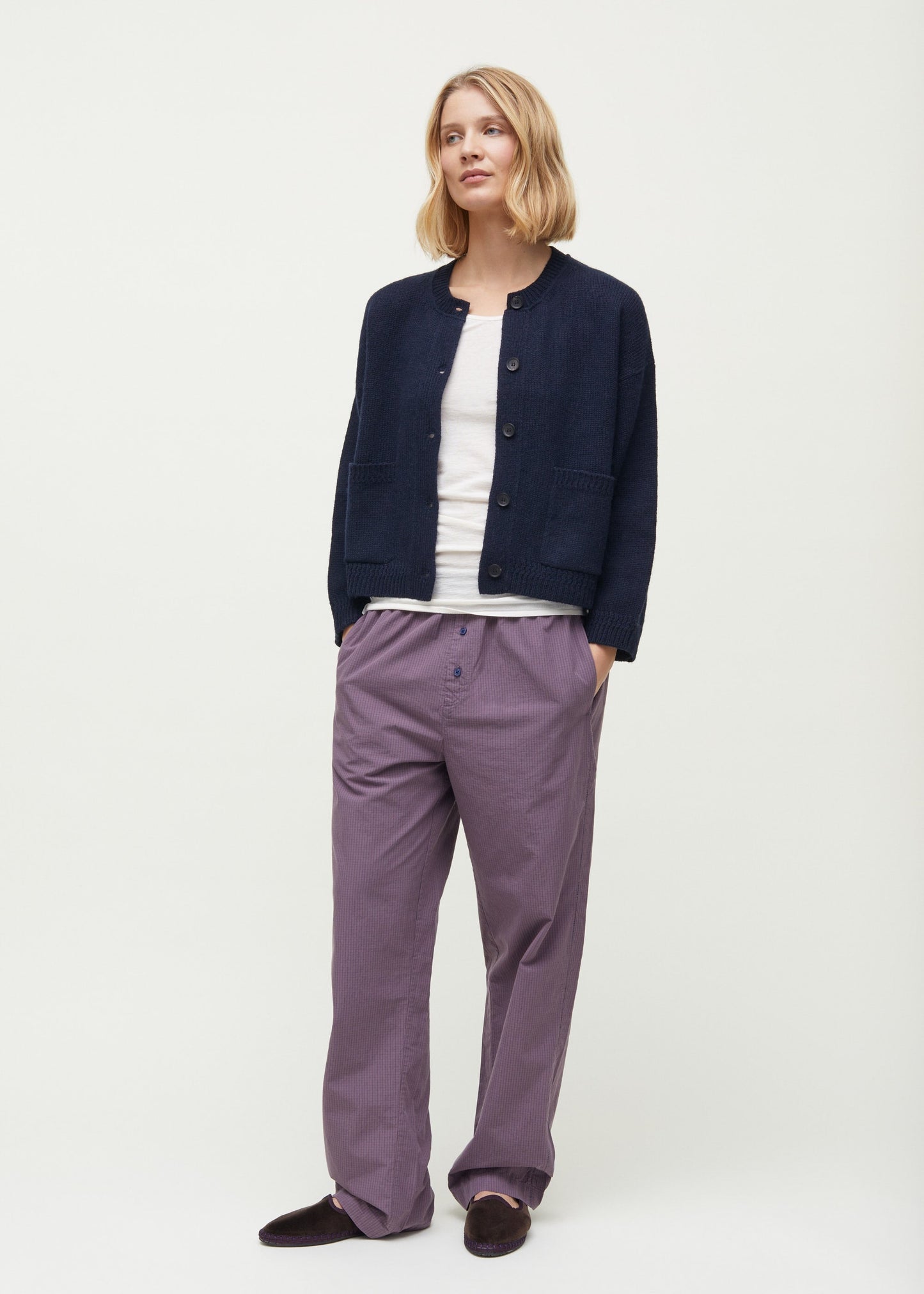 A woman stands with her hands in her pockets, donning a black button-up Cobe Cardigan from Aiayu over a white shirt and loose-fitting purple pants. She has shoulder-length blonde hair and a neutral facial expression.