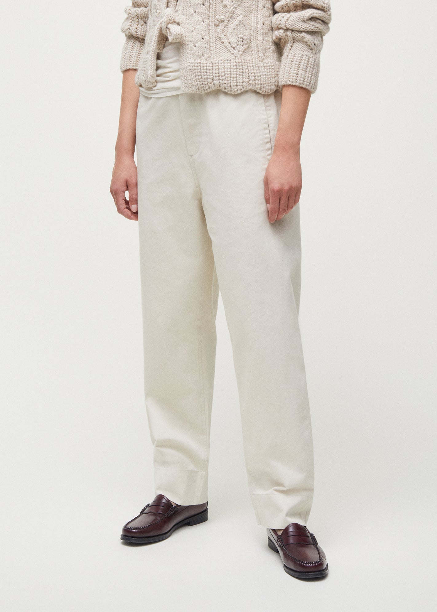 A person is wearing a beige knit sweater, the Coco Pant Twill high-waisted white pants from Aiayu in a classic straight fit, and brown loafers, standing against a plain white background.