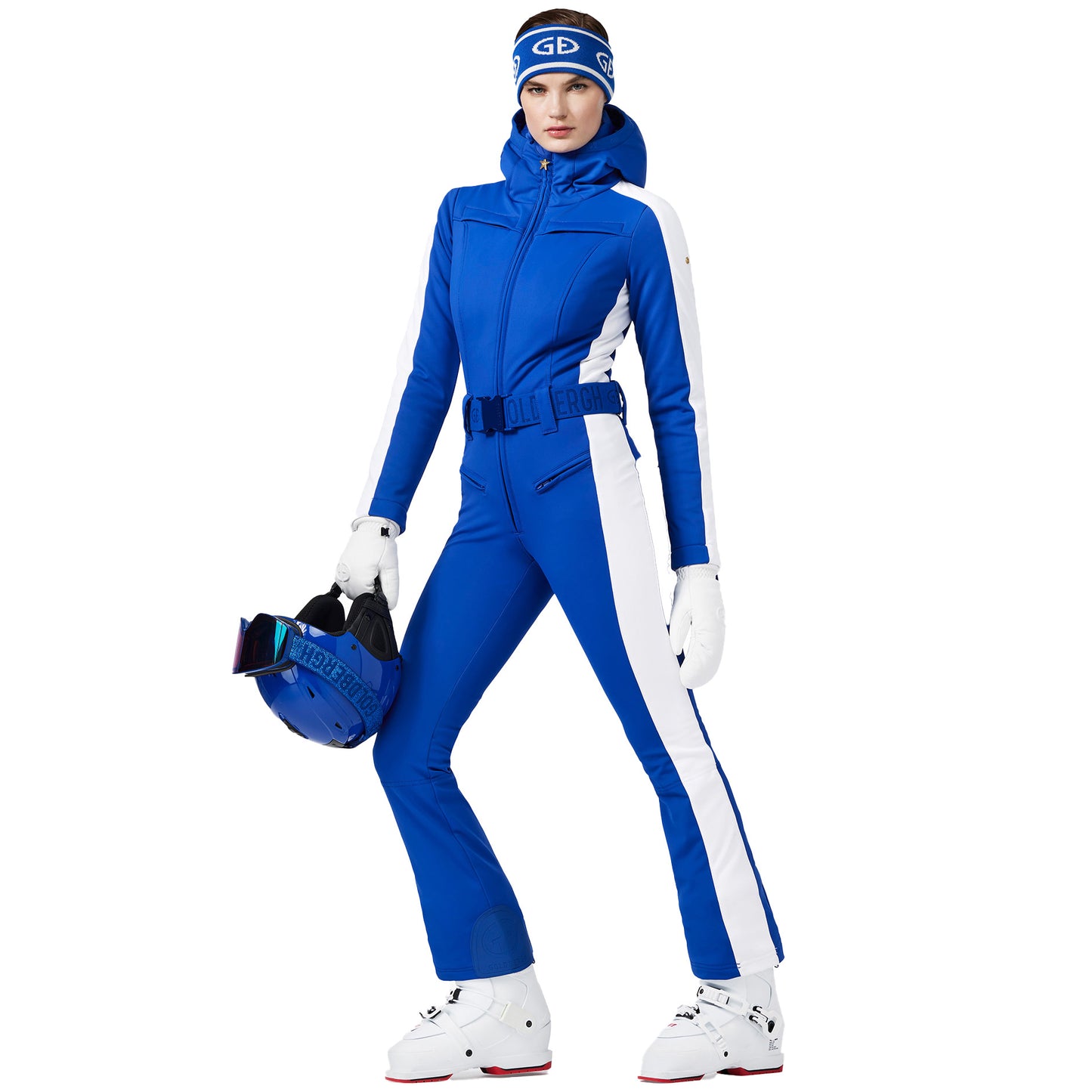 A person wearing the Coureur Ski Suit in Dazzling Blue by Goldbergh, along with a coordinating beanie and holding a helmet, stands against a white background.