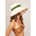 A woman in a ruched tan top and large white floppy sun protection hat adorned with a green stripe, with her face turned away from the camera, wearing the "Dia" natural/terracotta hat from Lola Hats.