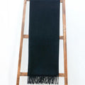 Alashan's The Classic Throw, a Mongolian cashmere black scarf with fringe, hangs over a wooden ladder against a white background.