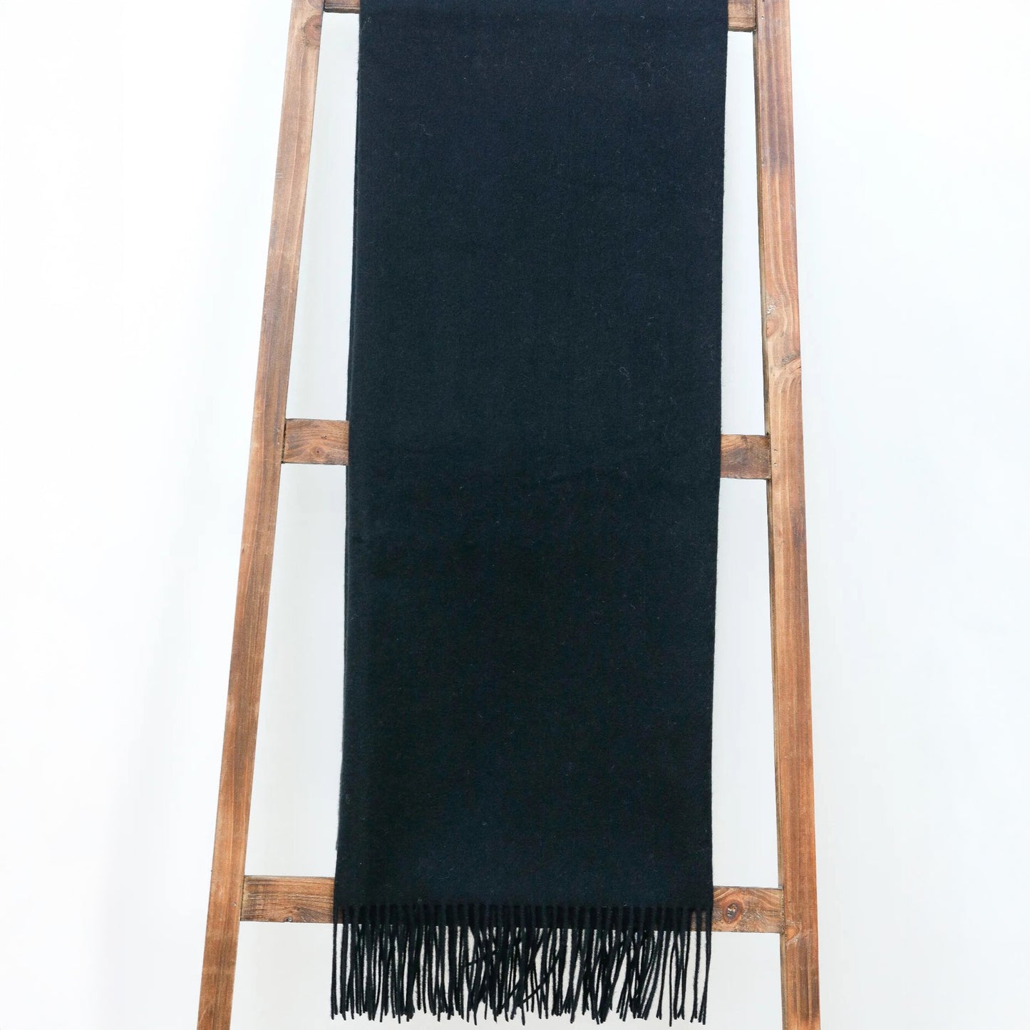 Alashan's The Classic Throw, a Mongolian cashmere black scarf with fringe, hangs over a wooden ladder against a white background.