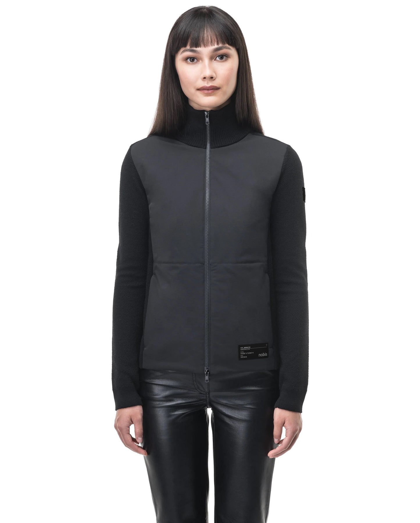 A person wearing the Evo Women's Performance Full Zip Sweater by Nobis, featuring stylish Merino wool trimming, pairs it with leather pants and stands against a white background.