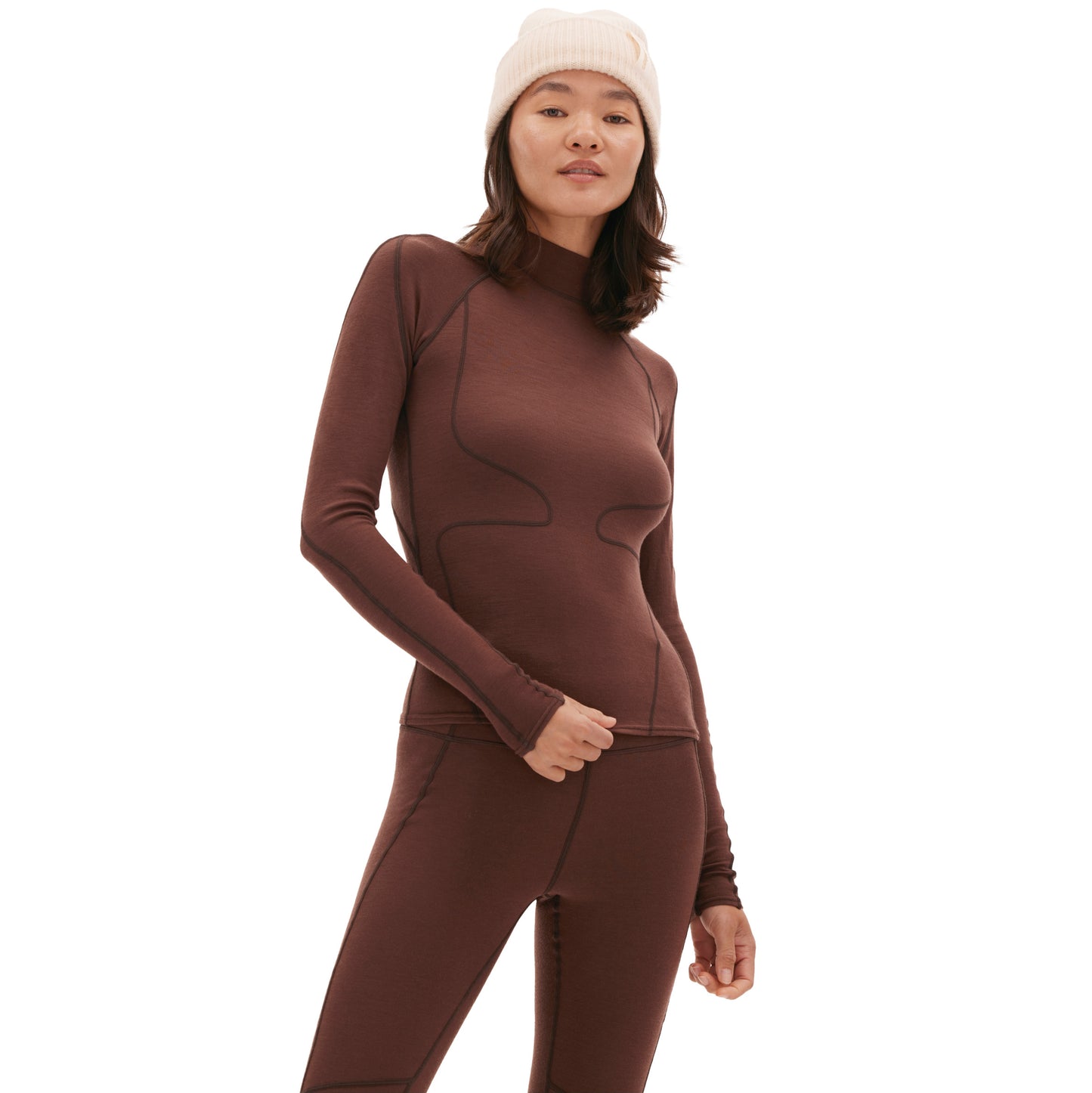 A person stands against a plain background, wearing the Fay Merino Top by Halfdays and brown leggings, complemented by a white beanie.