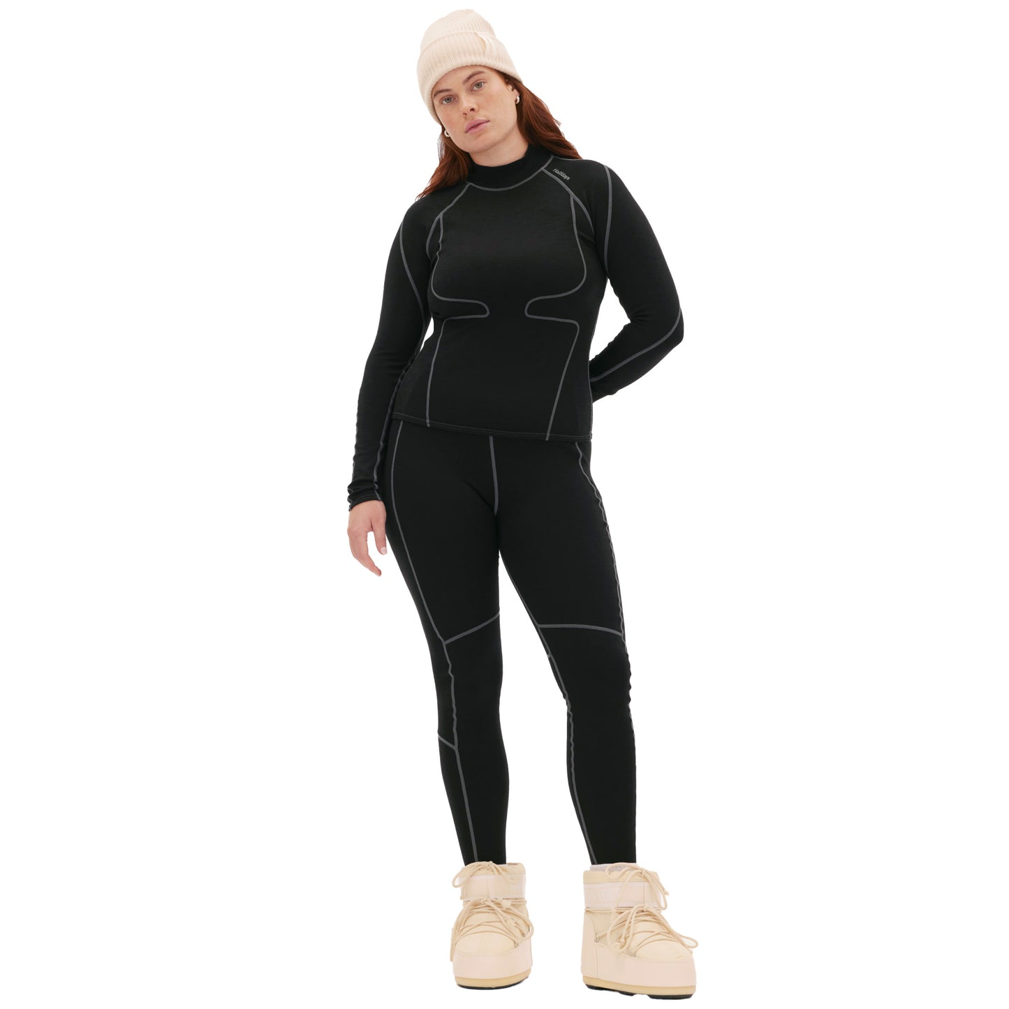 Person wearing the Fay Merino Top by Halfdays, a sleek black athletic outfit with white stitching, complemented by a cozy merino wool beanie and beige boots, stands against a white background.