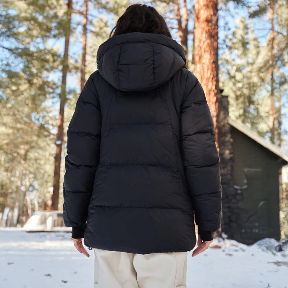 Wearing the Tabei Parka by Halfdays, a person stands in a snowy, wooded area with a small cabin in the background, embodying the tranquility of winter days.