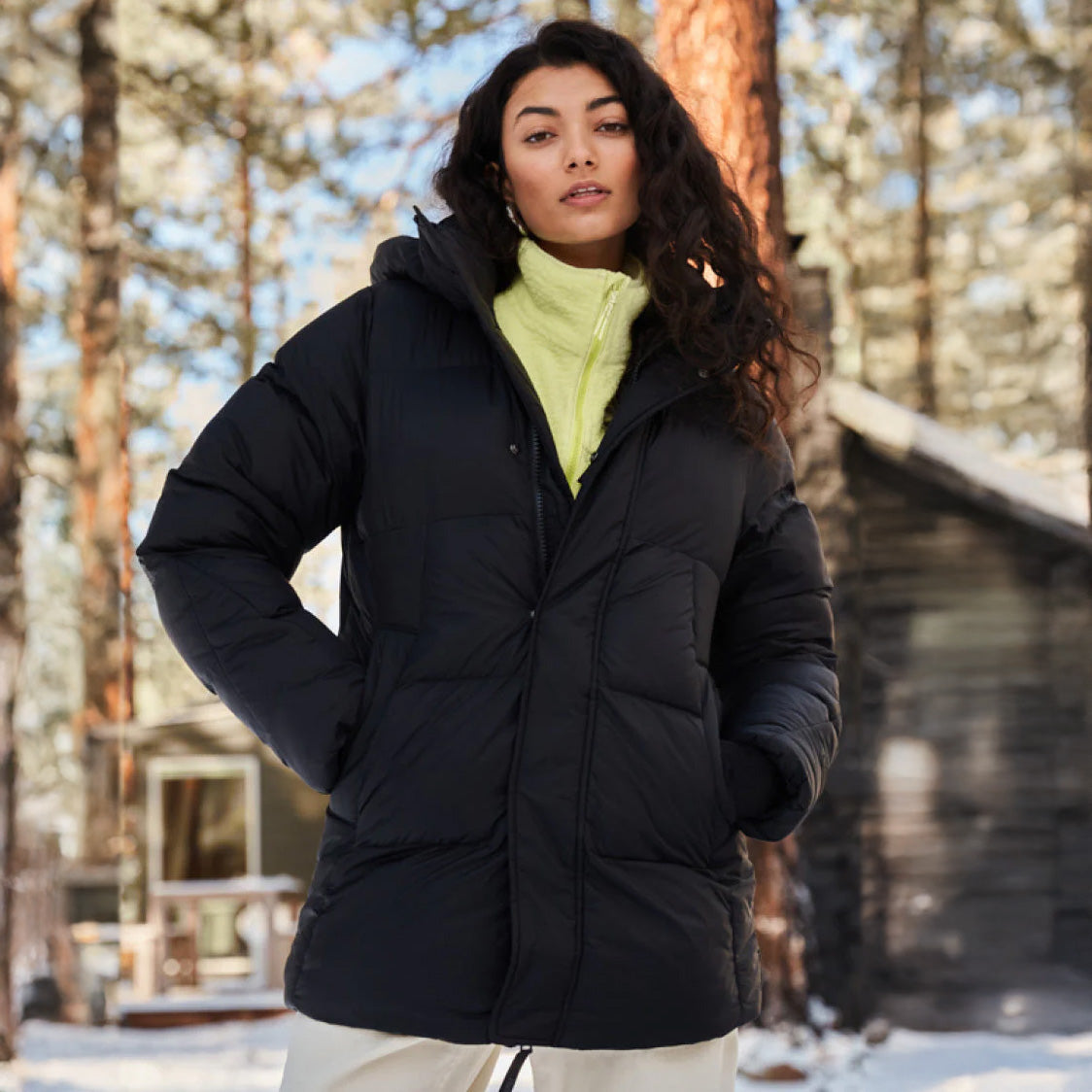 Wearing a Halfdays Tabei Parka, a person stands outdoors amidst the serene embrace of snow-covered trees near a wooden cabin on a wintry day.