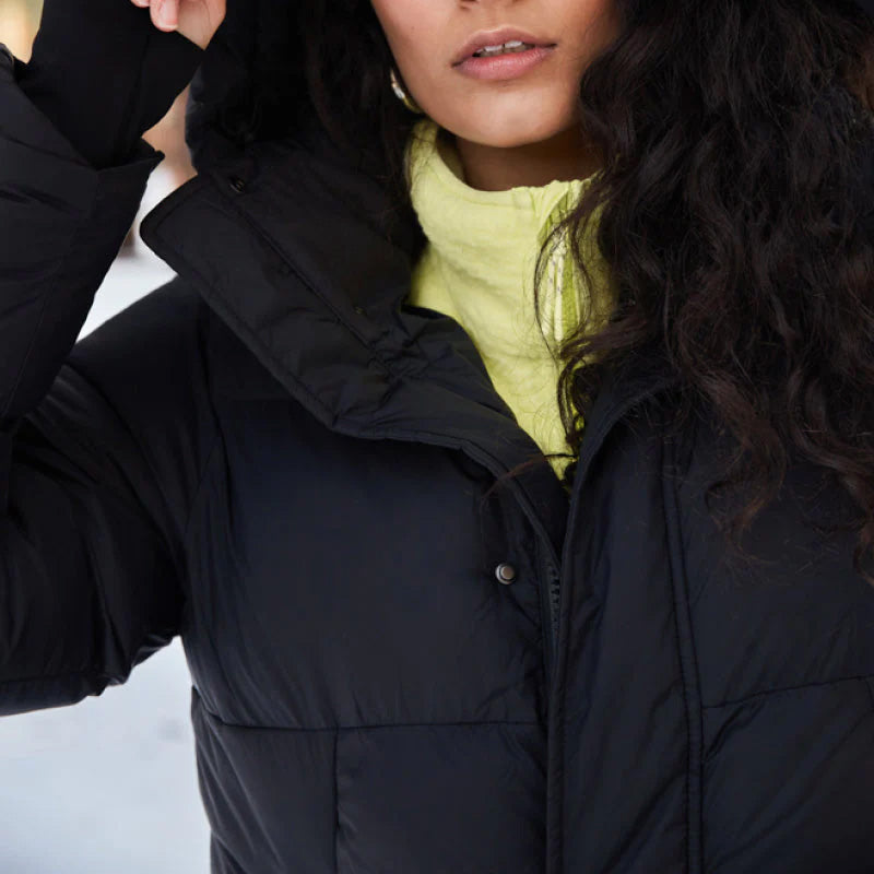 A person adjusts the hood of a Tabei Parka by Halfdays, wearing it over a yellow top to brace against snowy winter days.