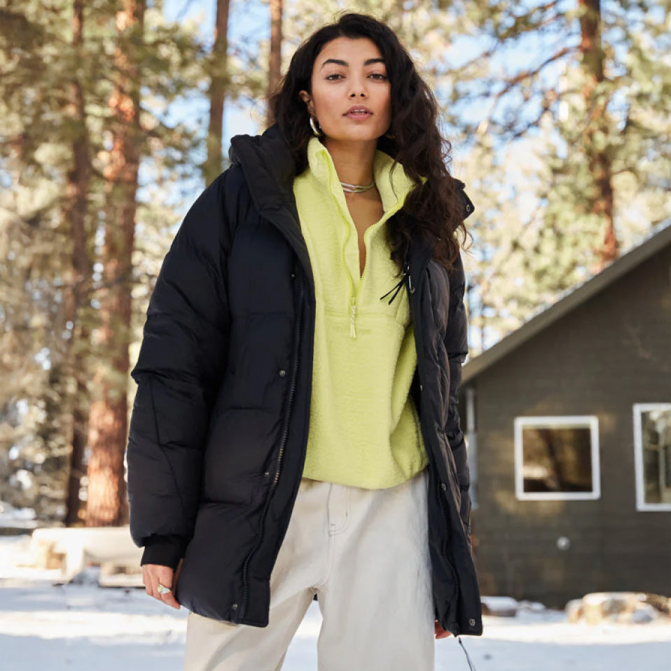 A person stands outside near a house, wrapped in the Halfdays Tabei Parka over a yellow top and light pants. Snow blankets the ground, capturing winter's serene essence with tall trees majestically in the background.