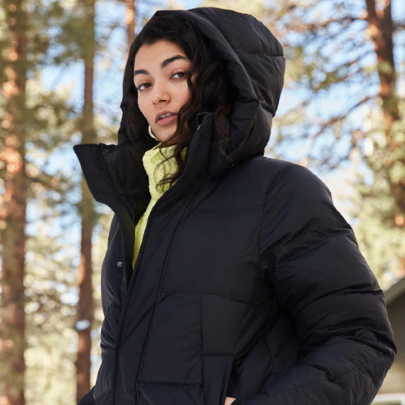 Wrapped warmly in the black Tabei Parka by Halfdays, a person stands outdoors with its insulated protection and hood, while trees sway gently in the background—a perfect shield against chilly winter days.