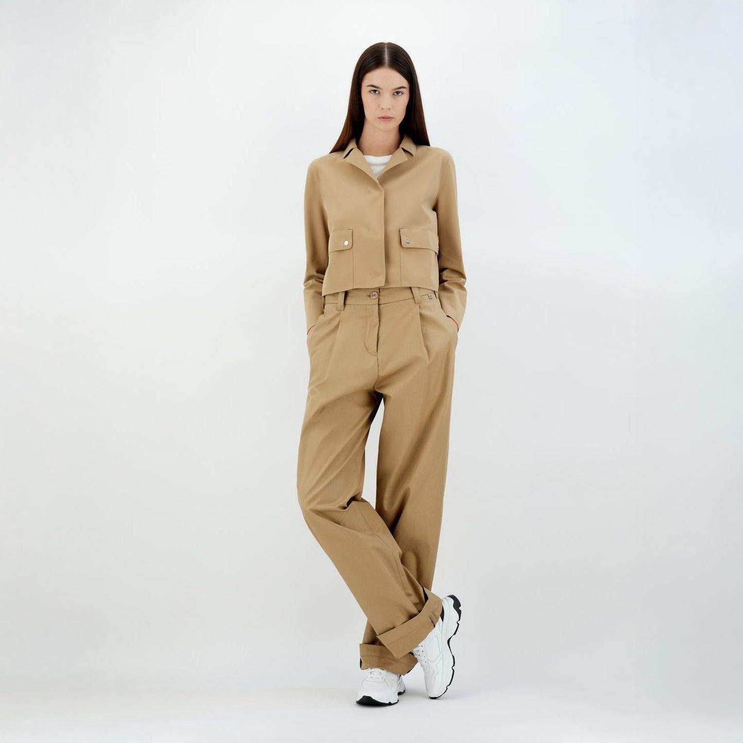 A person stands against a plain background with hands in pockets, wearing the Herno W Scuba Short Jacket Patch Pockets in camel and matching pants from Sensitive® Fabrics, paired with white sneakers.