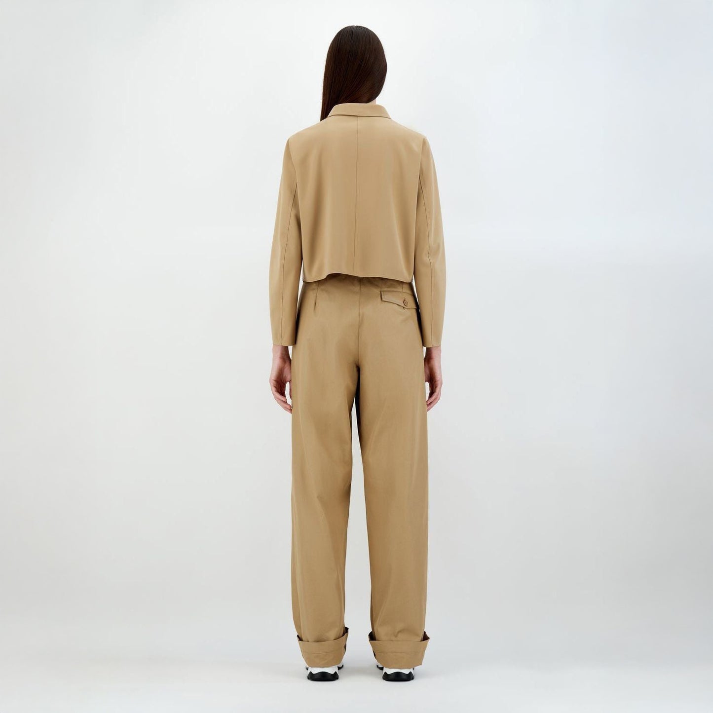 A person with long hair wears the Herno W Scuba Short Jacket Patch Pockets in Camel, featuring wide-legged pants and sneakers, standing against a plain white background.