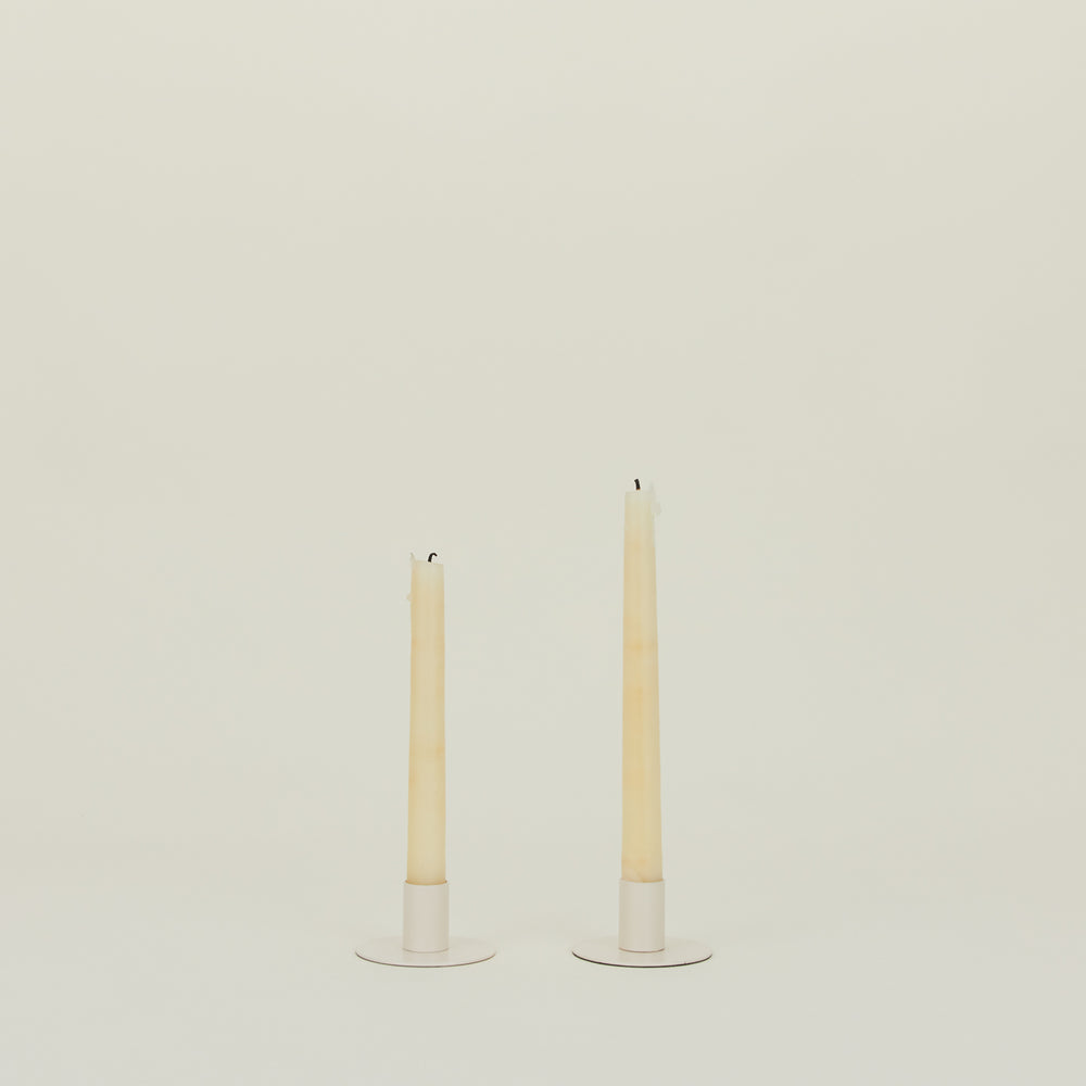 Two tall, unlit taper candles elegantly rest on Hawkins New York's Essential Metal Candle Holder set in ivory, made of powder-coated iron, against an off-white backdrop.