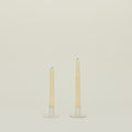 Two tall, unlit taper candles elegantly rest on Hawkins New York's Essential Metal Candle Holder set in ivory, made of powder-coated iron, against an off-white backdrop.