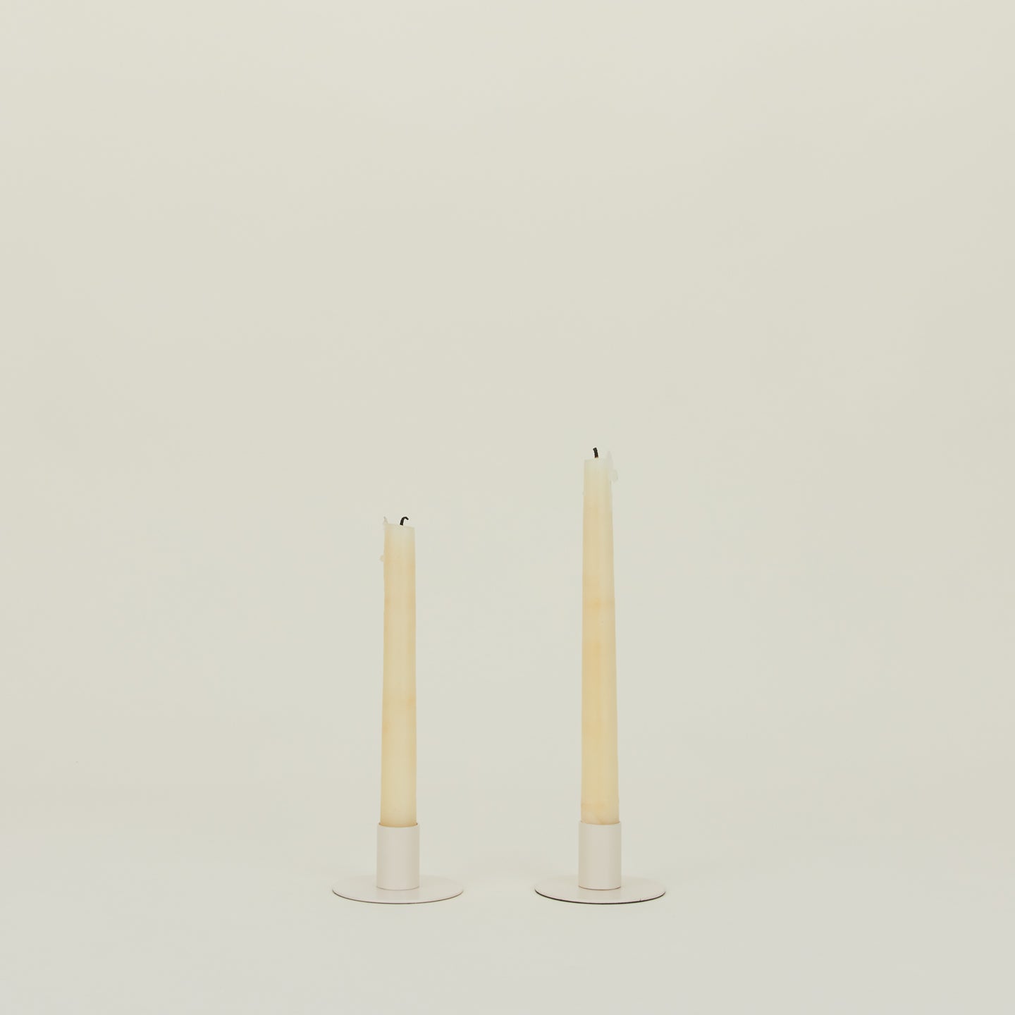 Two tall, unlit taper candles elegantly rest on Hawkins New York's Essential Metal Candle Holder set in ivory, made of powder-coated iron, against an off-white backdrop.