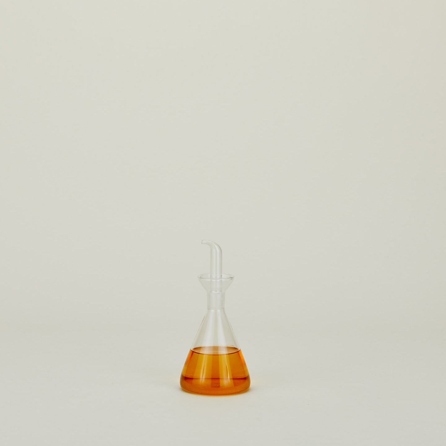 Part of Hawkins New York's Essential Collection, the clear glass Medium Pyramid Kitchen Bottle with amber liquid stands elegantly against a plain light background, blending scientific charm with easy living home basics.