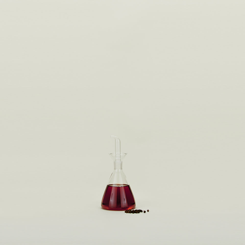 A Medium Pyramid glass bottle from the Essential Kitchen Bottles collection by Hawkins New York, half-filled with red liquid, sits on a light surface next to a small pile of black peppercorns.