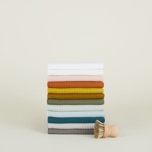 Next to a wooden brush with bristles against a plain background sits a set of colorful Essential Waffle Dish Towels by Hawkins New York. These super absorbent towels are quick to dry and perfect for any kitchen space.