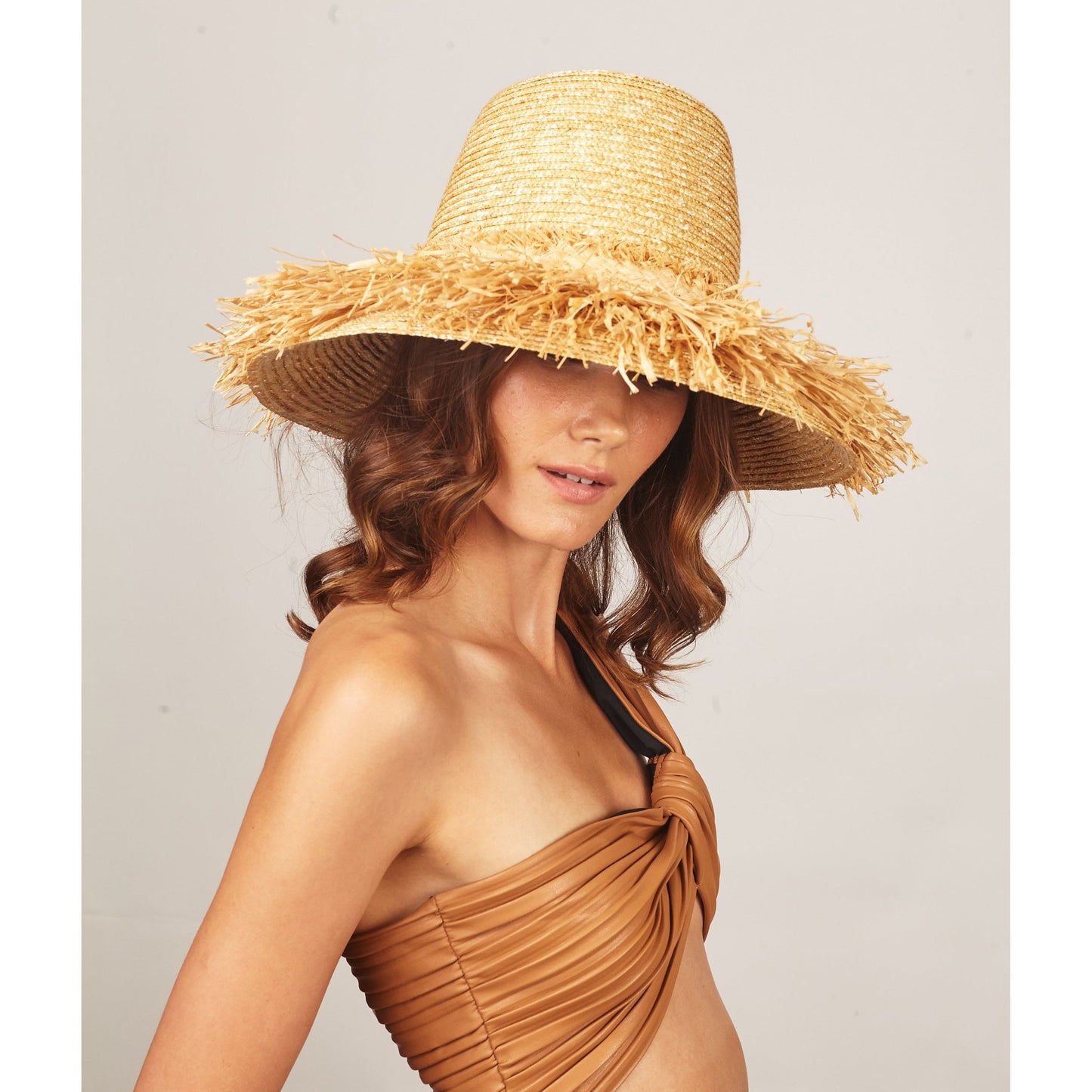 A woman wearing Lola Hats' wide-brimmed wheat straw hat and a brown swimsuit poses against a neutral background.