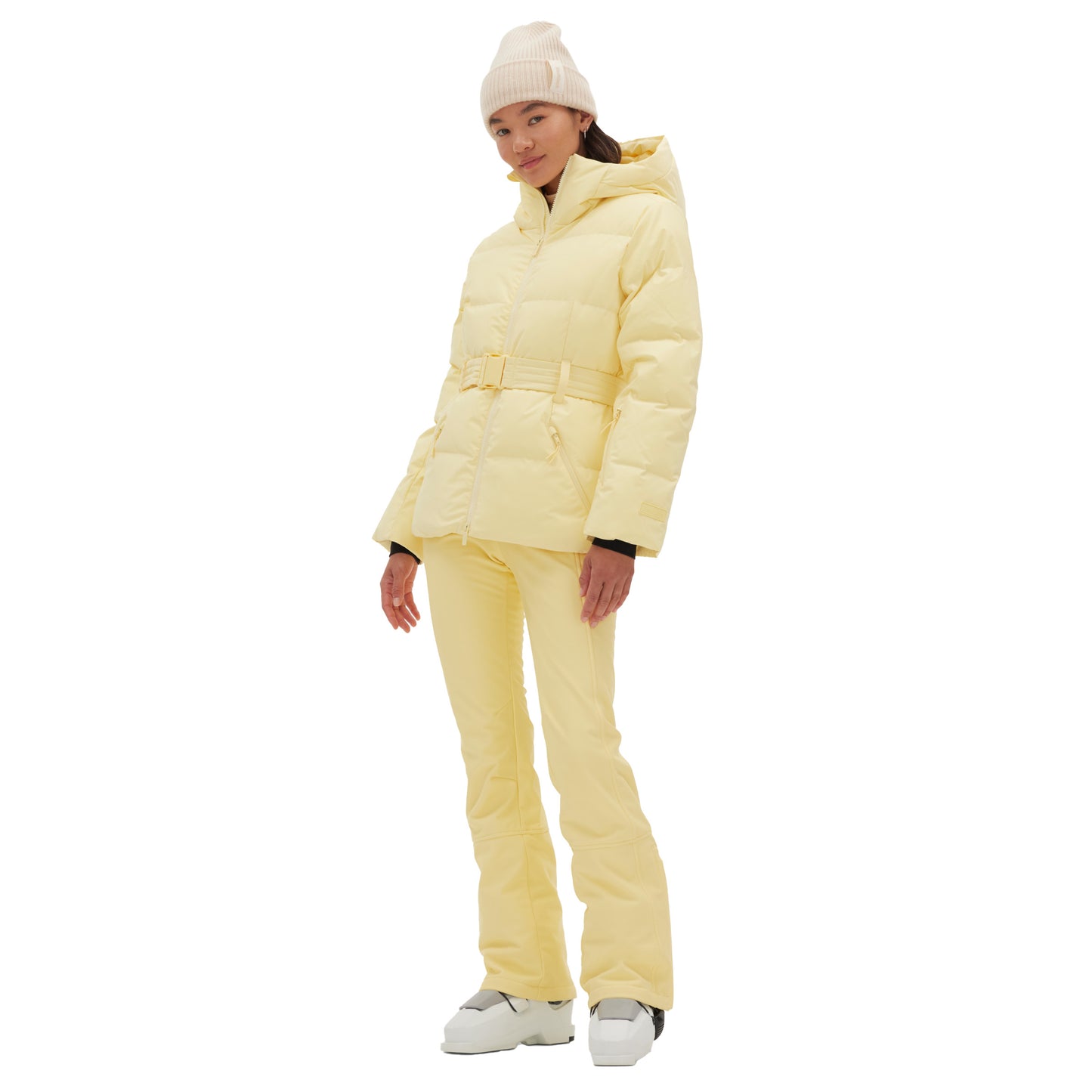 Dressed in a Halfdays Hedy Puffer Jacket, complemented by matching pants, a beige beanie, and white boots, an individual stands against a white background, embodying mountain performance features perfect for the slopes.