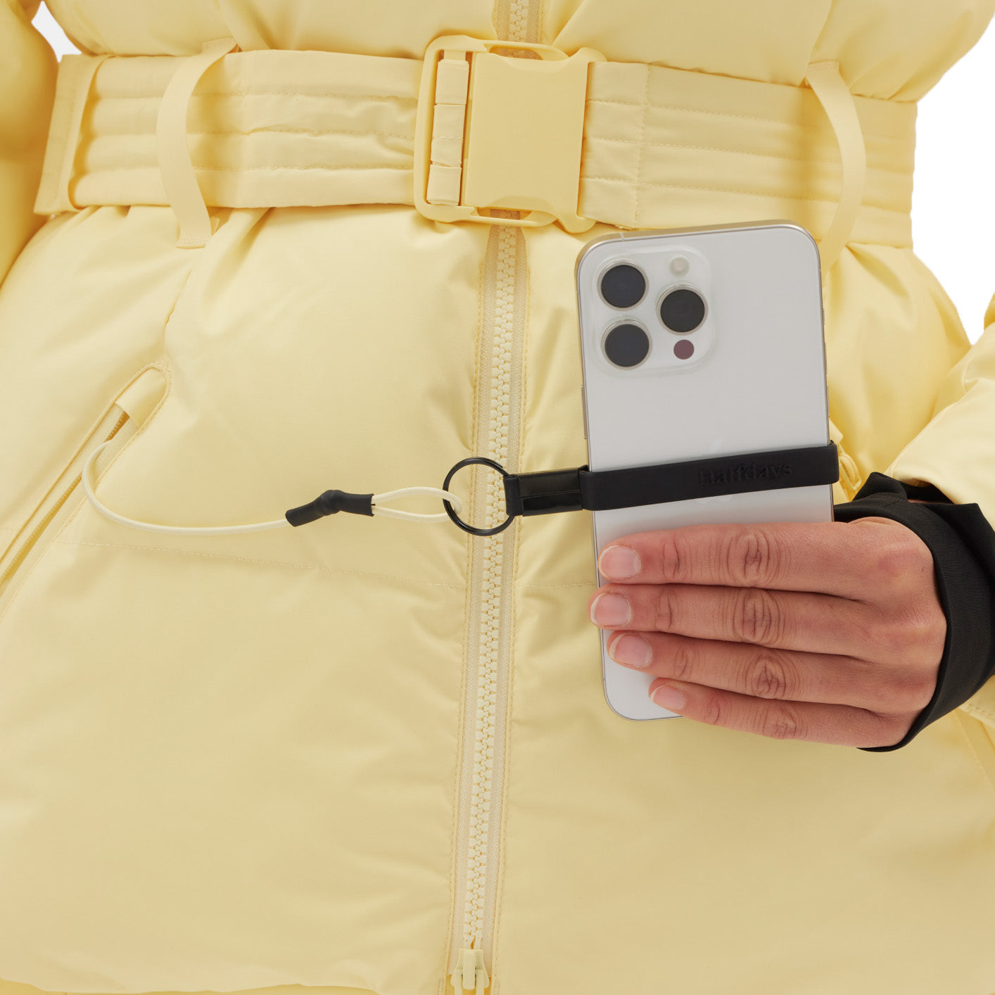 A person in a yellow Hedy Puffer Jacket from Halfdays holds a smartphone with a black strap and lanyard connected to a zipper pull, ready for the slopes.