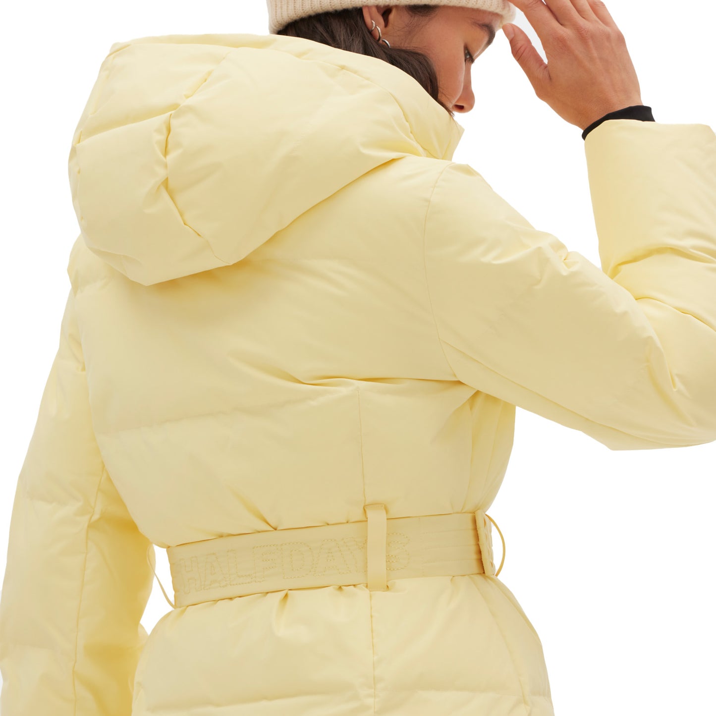 A person wearing the Hedy Puffer Jacket by Halfdays in a pale yellow color, along with a beige beanie, adjusts the hood for enhanced mountain performance features.