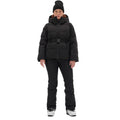 A person stands facing forward wearing a black Hedy Puffer Jacket by Halfdays, complemented by black pants and white boots. They sport a black beanie and belt, great for hitting the slopes. The jacket hood is crafted with mountain performance features.