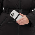 A person wearing the Hedy Puffer Jacket by Halfdays holds a smartphone with a black strap and lanyard attachment, ready to capture moments on the slopes.