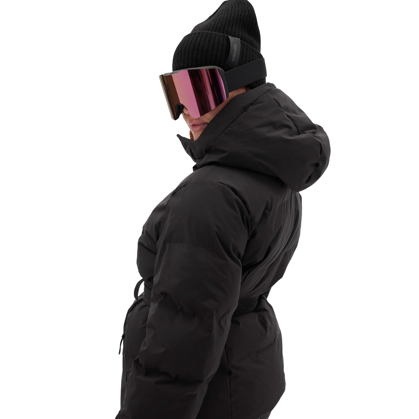 A person dressed in a black Hedy Puffer Jacket by Halfdays, paired with a black beanie and reflective ski goggles, glances to the side against a plain white background, poised for the slopes with mountain performance features.