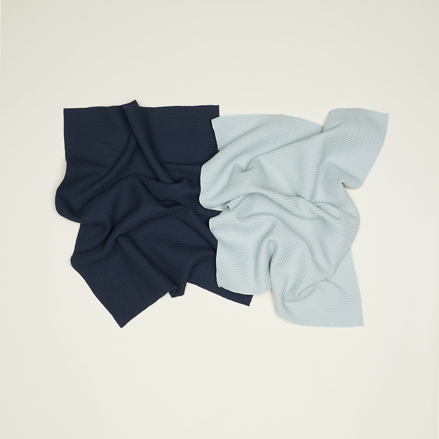 Two crumpled fabric squares lie side by side on a light surface: one dark blue and one light gray, both are super absorbent waffle weave kitchen towels from the Hawkins New York Essential Waffle Dish Towel set.