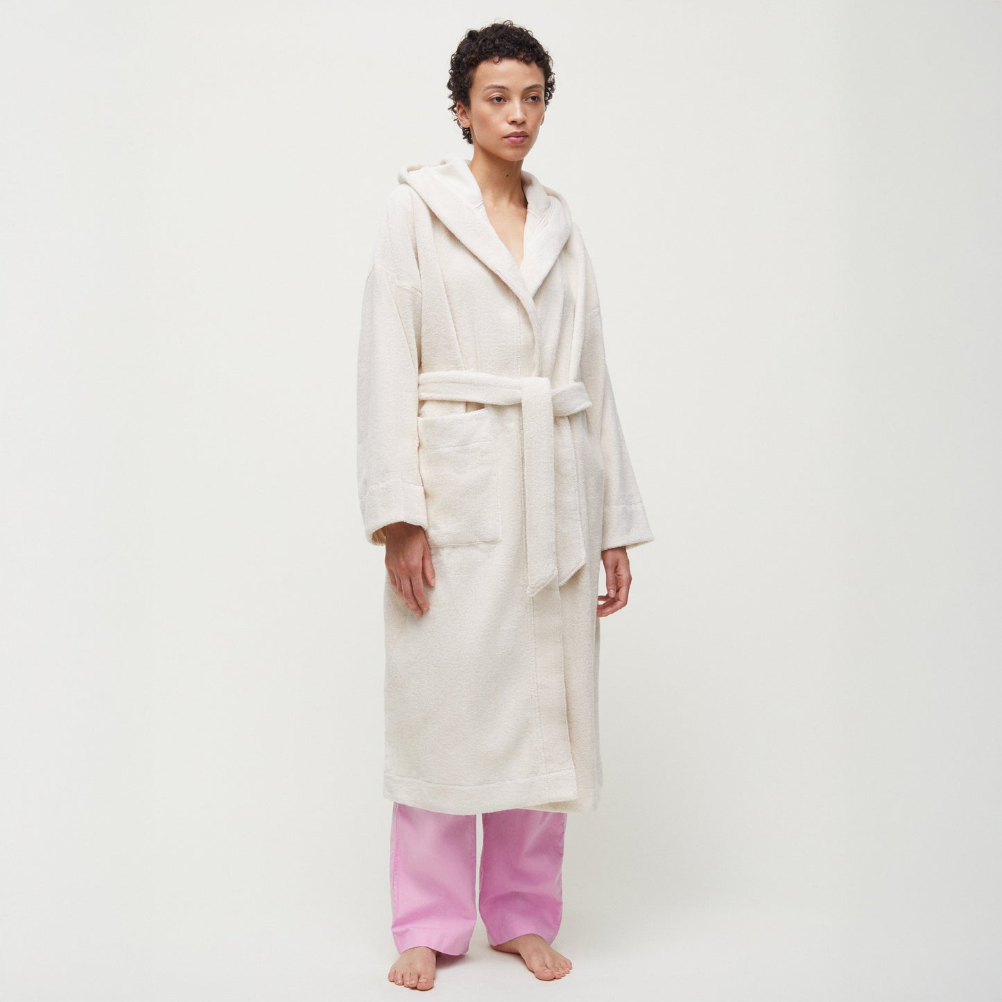 Barefoot against a simple backdrop, a person wears Aiayu's organic Chetna cotton Hooded Terry Bathrobe in white and pink pants, capturing the elegance and simplicity of traditional India.