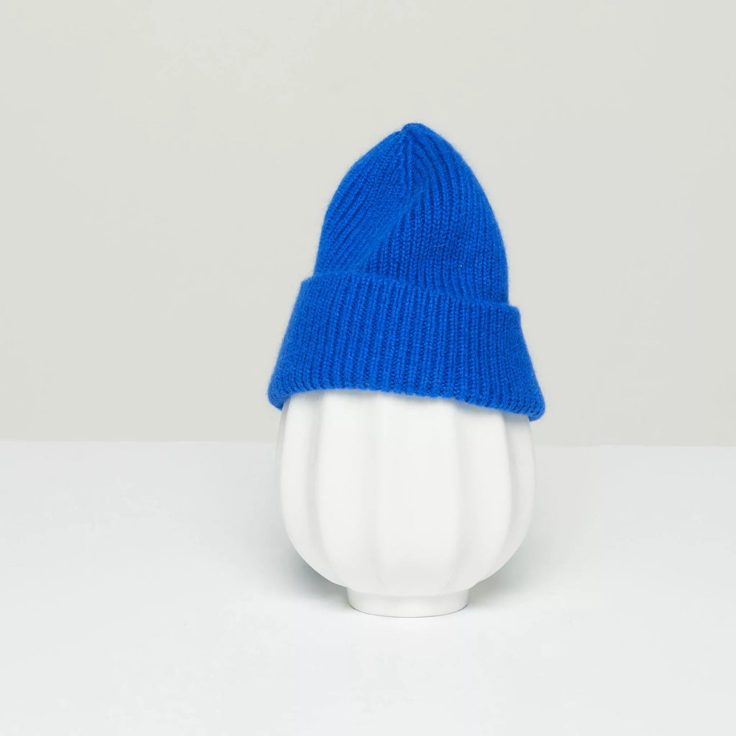 A Royal Azure K Beanie by Le Bonnet, made from luxurious lambswool, is displayed on a white ribbed vase against a light gray background.