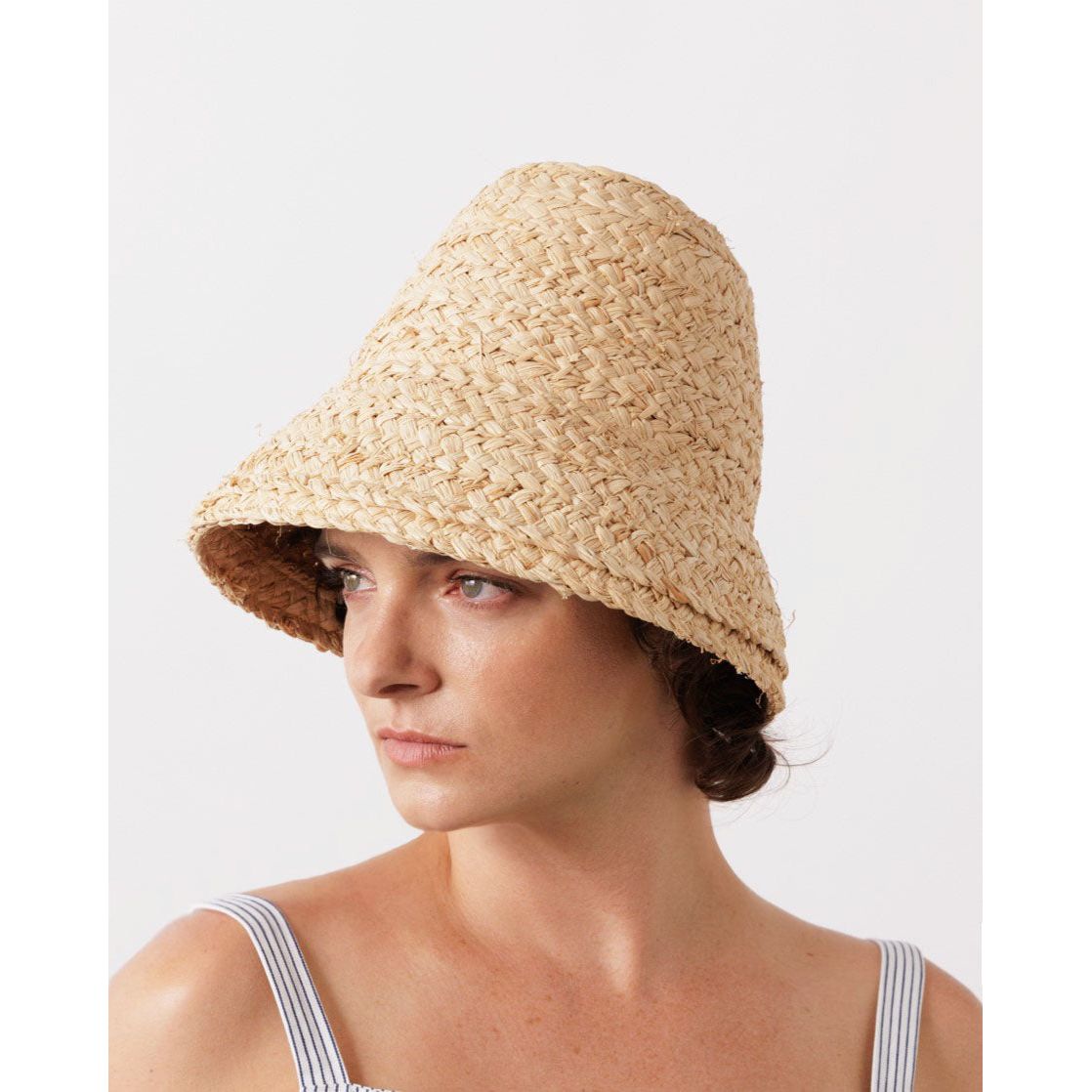 A person with brown hair is wearing a Petit Nap Hat, Natural by Lola Hats woven from natural raffia braid and a striped top, looking to the left against a plain background.