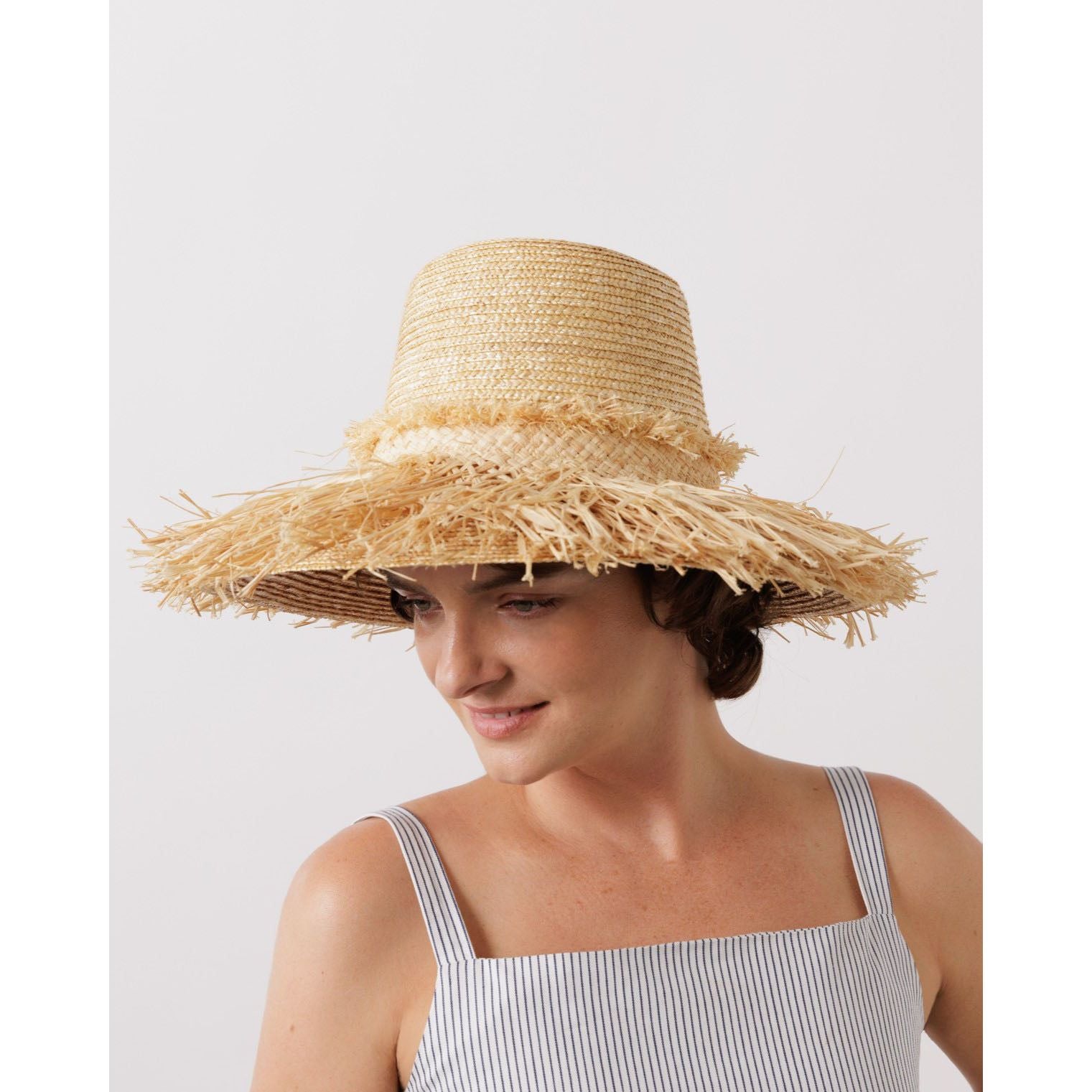 A woman wearing a Lola Hats Hula Skirt made of natural wheat straw with frayed edges, dressed in a striped outfit, looks downward with a subtle smile.