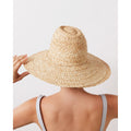 A person seen from behind wearing a wide-brimmed raffia hat (Fiscolo, Natural) by Lola Hats, with a hint of striped clothing visible on the shoulders.