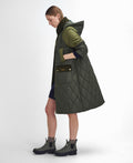 A person with light brown hair is standing sideways, wearing a long, dark green Barbour W Cookston Longline Quilted Jacket featuring diamond quilting and a detachable hood, paired with a blue dress and green ankle boots.