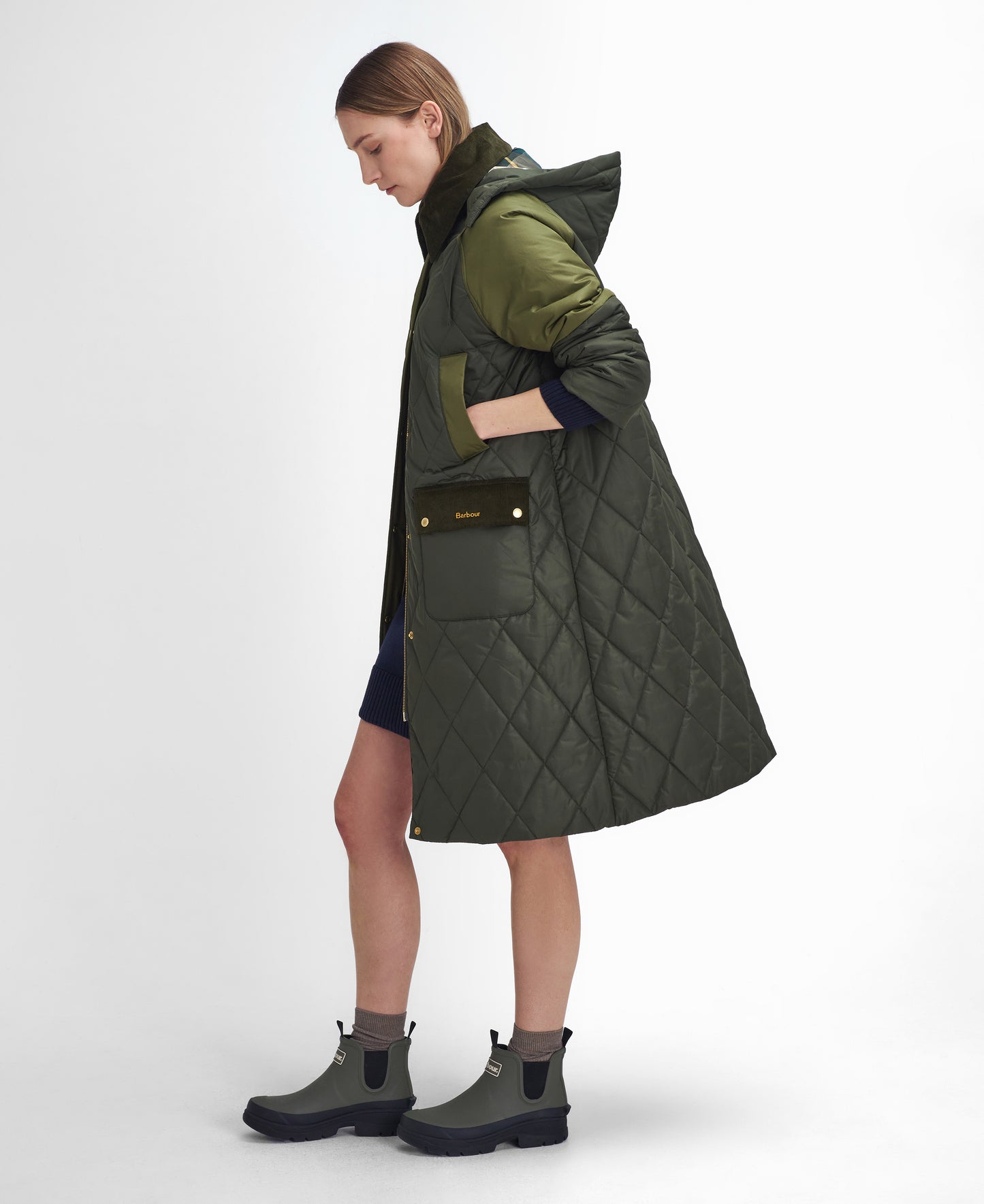 A person with light brown hair is standing sideways, wearing a long, dark green Barbour W Cookston Longline Quilted Jacket featuring diamond quilting and a detachable hood, paired with a blue dress and green ankle boots.