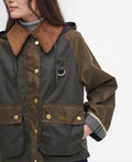 A woman dressed in a Barbour Reighton Spey Waxed Jacket with an oversized cord collar stands against a plain background. The visible parts include her torso and part of her head. The jacket, enhanced with gold buttons and large pockets, also boasts subtle tartan trims.