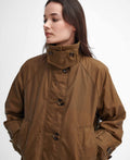 A person with dark hair wearing a brown buttoned jacket looks to the side with hands in pockets against a plain background, showcasing the perfect example of seasonal layering with the Barbour Hartwick Waxed Car Coat.