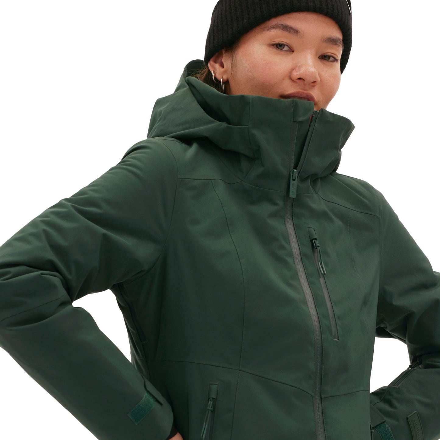 A woman wears a dark green Lawrence Jacket by Halfdays and a black beanie, standing with hands on hips against a plain backdrop.