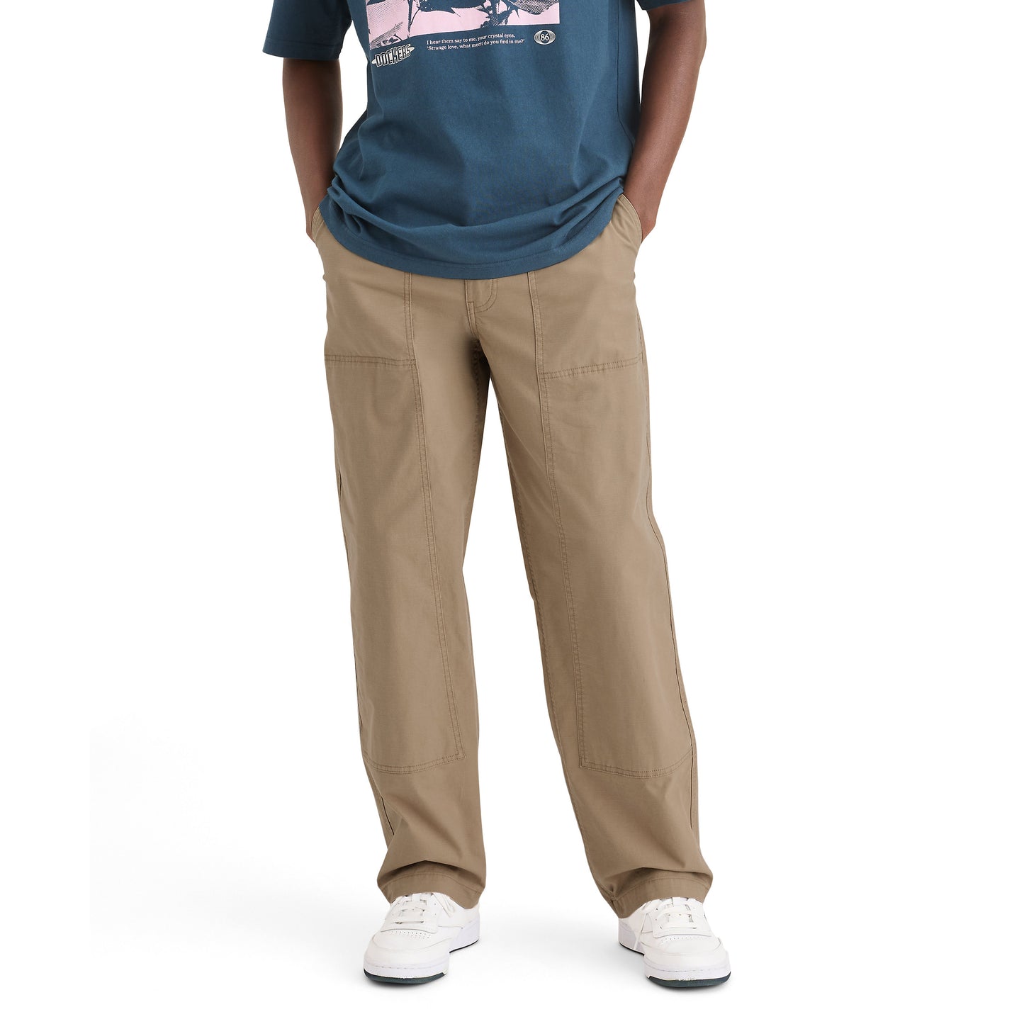 A person stands against a white background, hands in pockets, wearing a blue graphic T-shirt, Dockers' Genderless 86 Utility Pant in vintage-style khaki, and white sneakers.