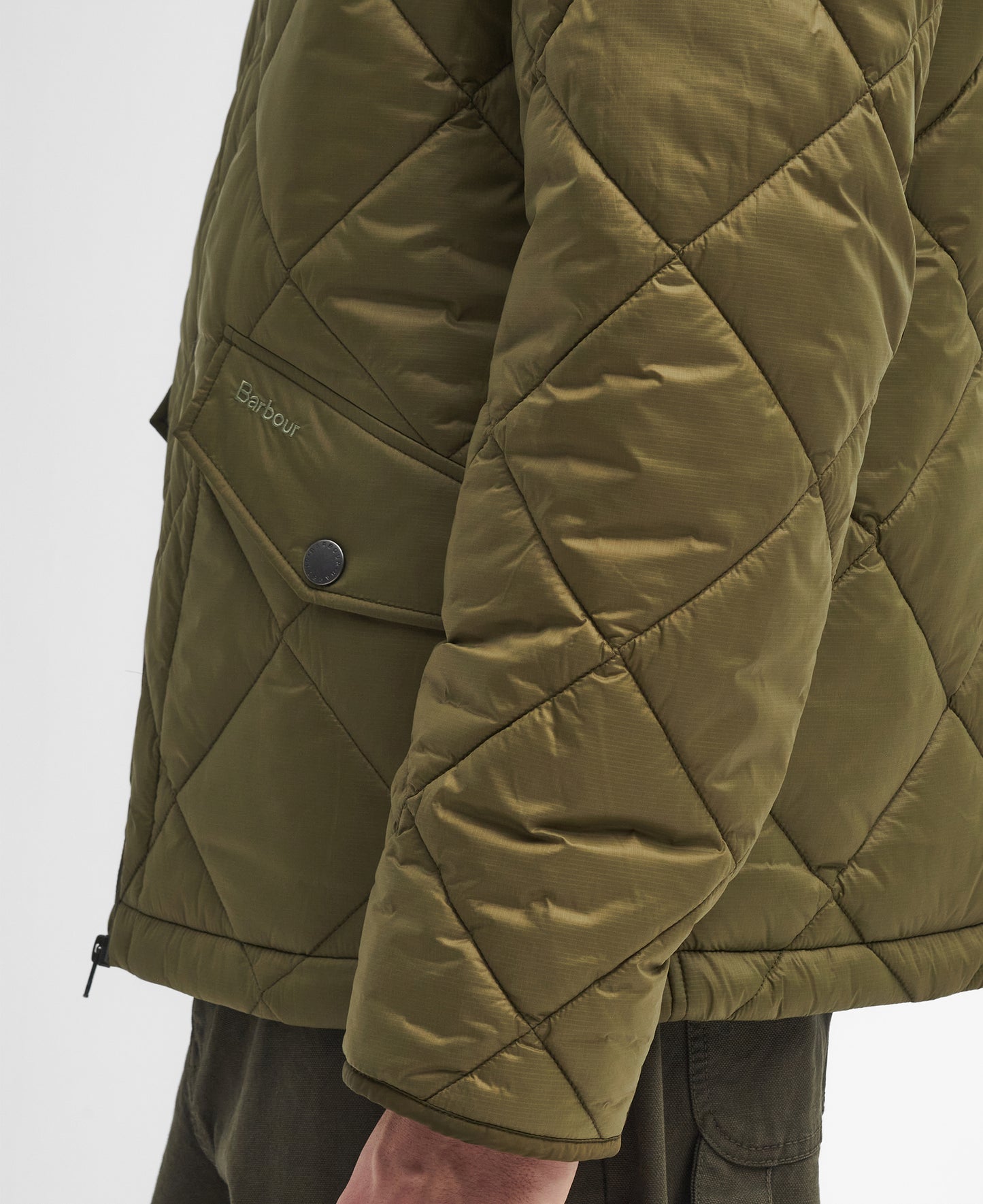 A close-up shot of a person wearing a Barbour Re-Engineered Endurance Quilted Jacket in green, highlighting the diamond quilting, distinct stitching, buttoned pockets, and the Barbour brand name on the pocket flap. The image captures only the torso and part of the arm.