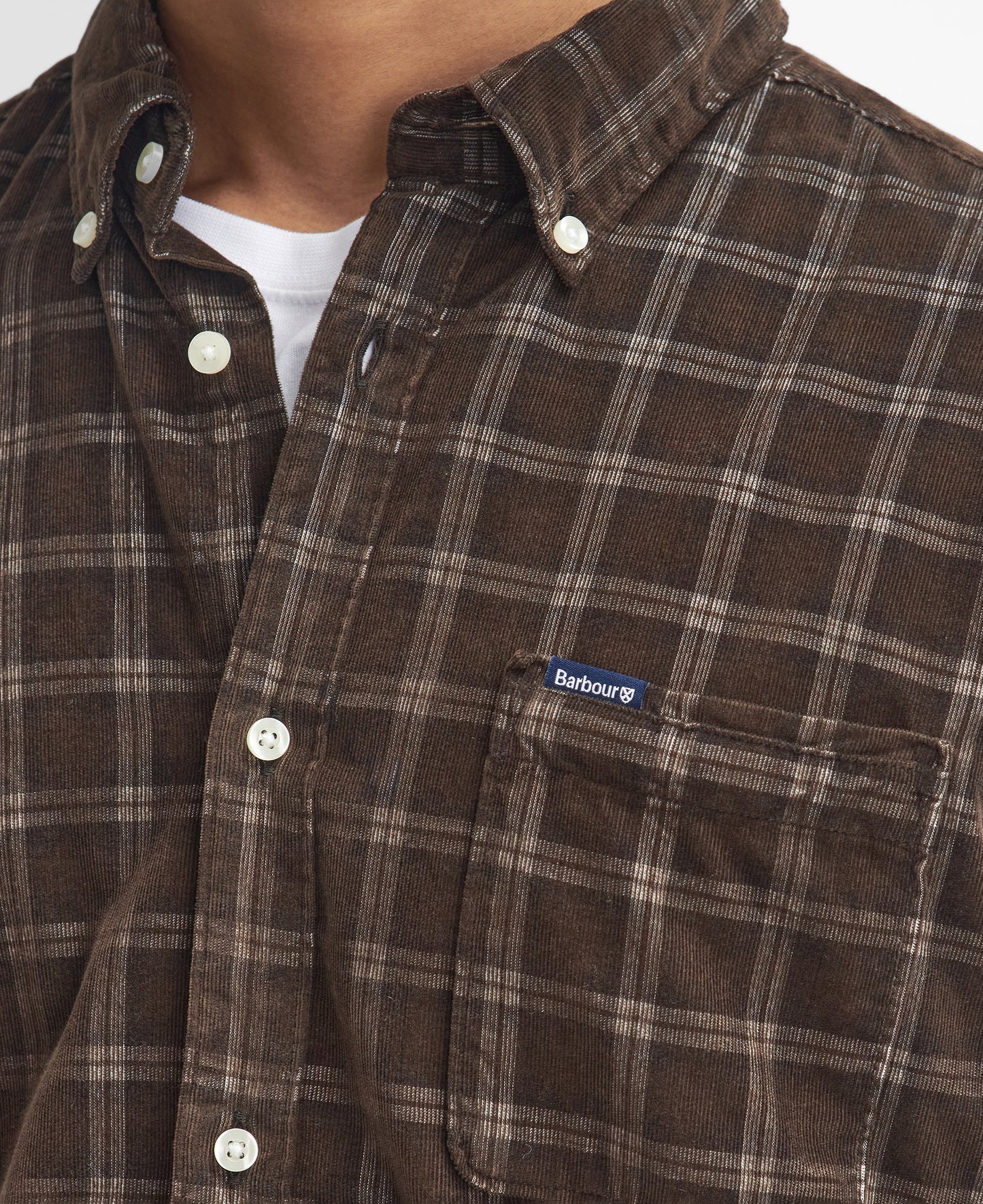 Close-up of a person wearing the Barbour Harthill Tailored Long Sleeved Cord Shirt, a brown plaid button-up featuring a small Barbour logo patch on the chest pocket, perfect for a smart-casual look.